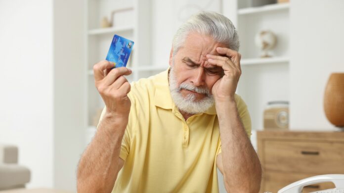 Descontos indevidos INSS: imagem retrata a angústia de aposentados afetados Idoso de expressão preocupada sentado em casa, segurando um cartão de crédito em uma mão e apoiando a outra na testa, representando tensão financeira.