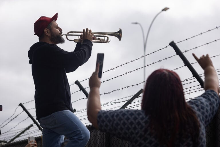Homem toca uma trombeta para celebrar a detenção pela Polícia Federal do Brasil, de forma preventiva neste sábado, o ex-presidente brasileiro Jair Bolsonaro, condenado por tentativa de golpe de Estado e quem foi encontrado na prisão domiciliar. Bolsonaro pretendia romper a barreira eletrônica que fiscalizava seus movimentos para fugir durante uma manifestação convocada em frente à sua casa por um de seus filhos | EFE/Vini Santa Rosa